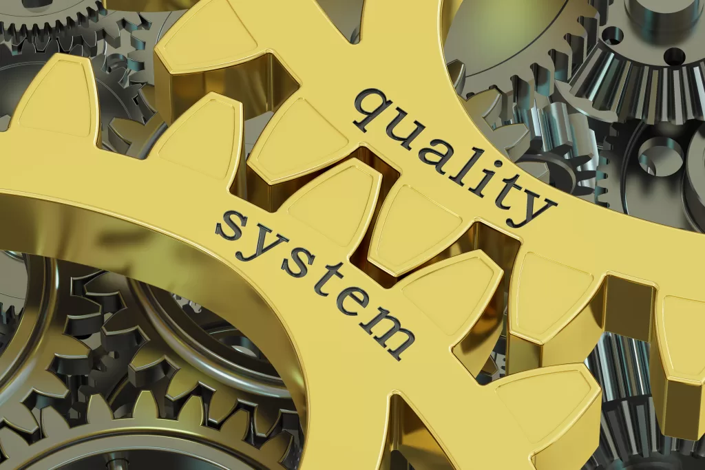 An image of 2 gold coloured cogs against each other, with the word quality one one cog and system on the other cog. This image represents Patient Guards Quality Assurance Medical Device Services.