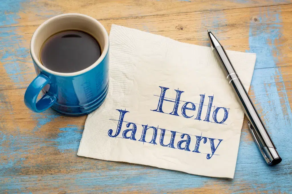 Napkin with Hello January written on it in Blue ink - There is a blue mug containing black coffee on a wooden table, and a silver/grey pen also on the table.
